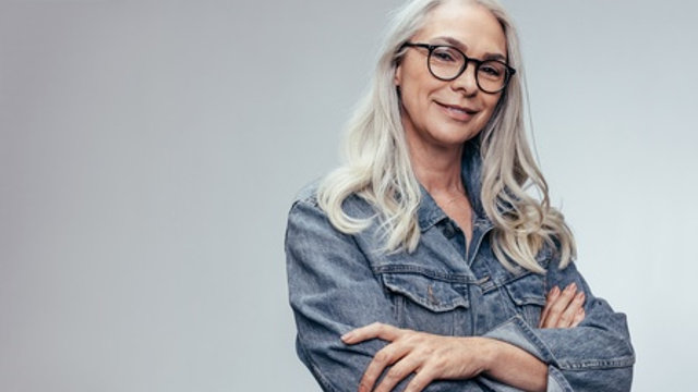 Beautiful mature caucasian female with eyeglasses looking at camera and smiling. Senior woman in casuals standing with her arms crossed against grey background.