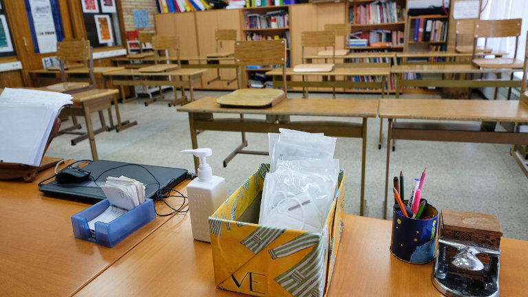 Mascarillas y gel desinfectante en la mesa del profesor de un aula donde se pueden observar los pupitres colocados y separados entre sí. EFE