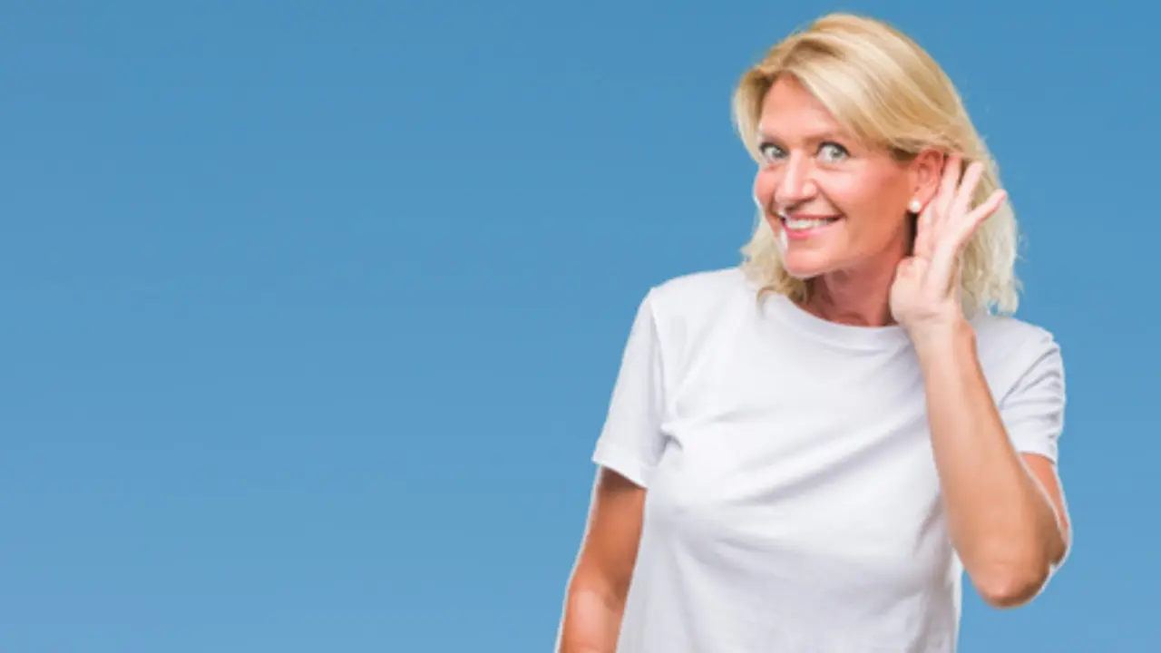 Middle age blonde woman over isolated background smiling with hand over ear listening an hearing to rumor or gossip. Deafness concept.