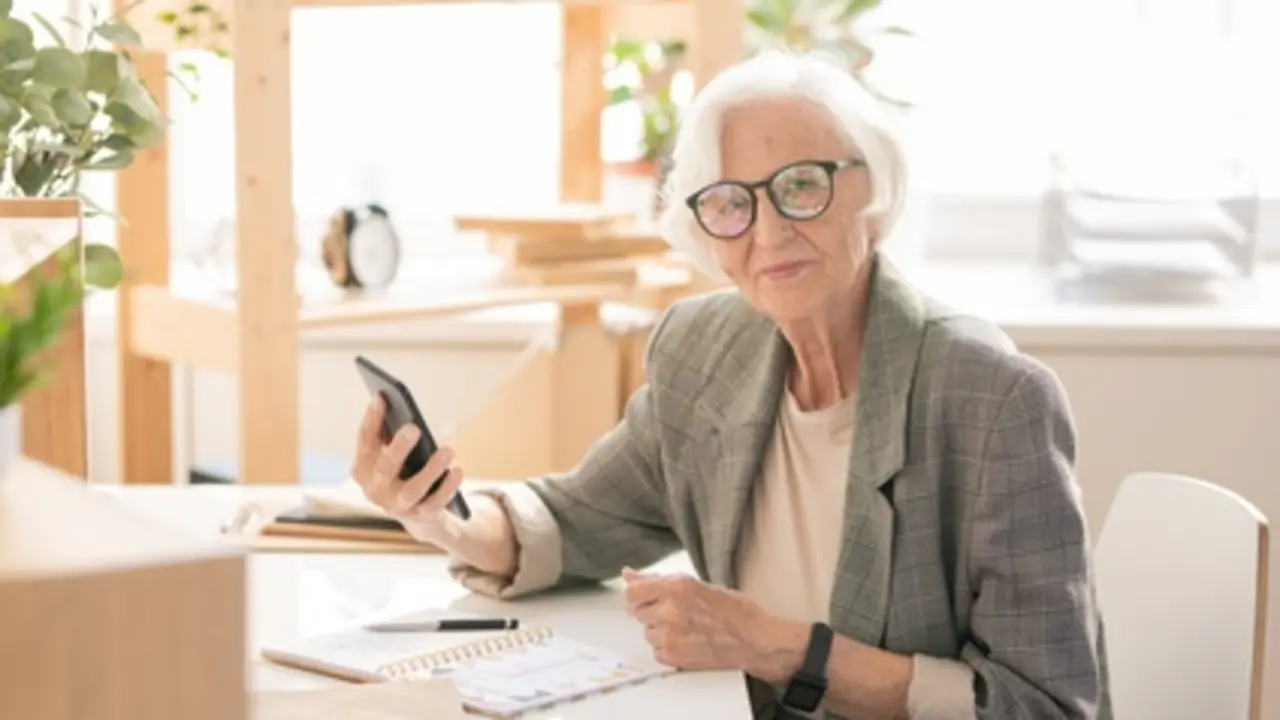 Modern and mobile senior female in formalwear and eyeglasses sitting by workplace in office in front of camera